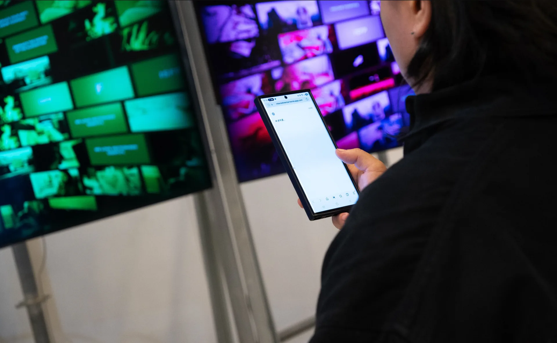 Visitor using a phone beside Ω screens showing changing images and text. Artwork by Jeanyoon Choi.