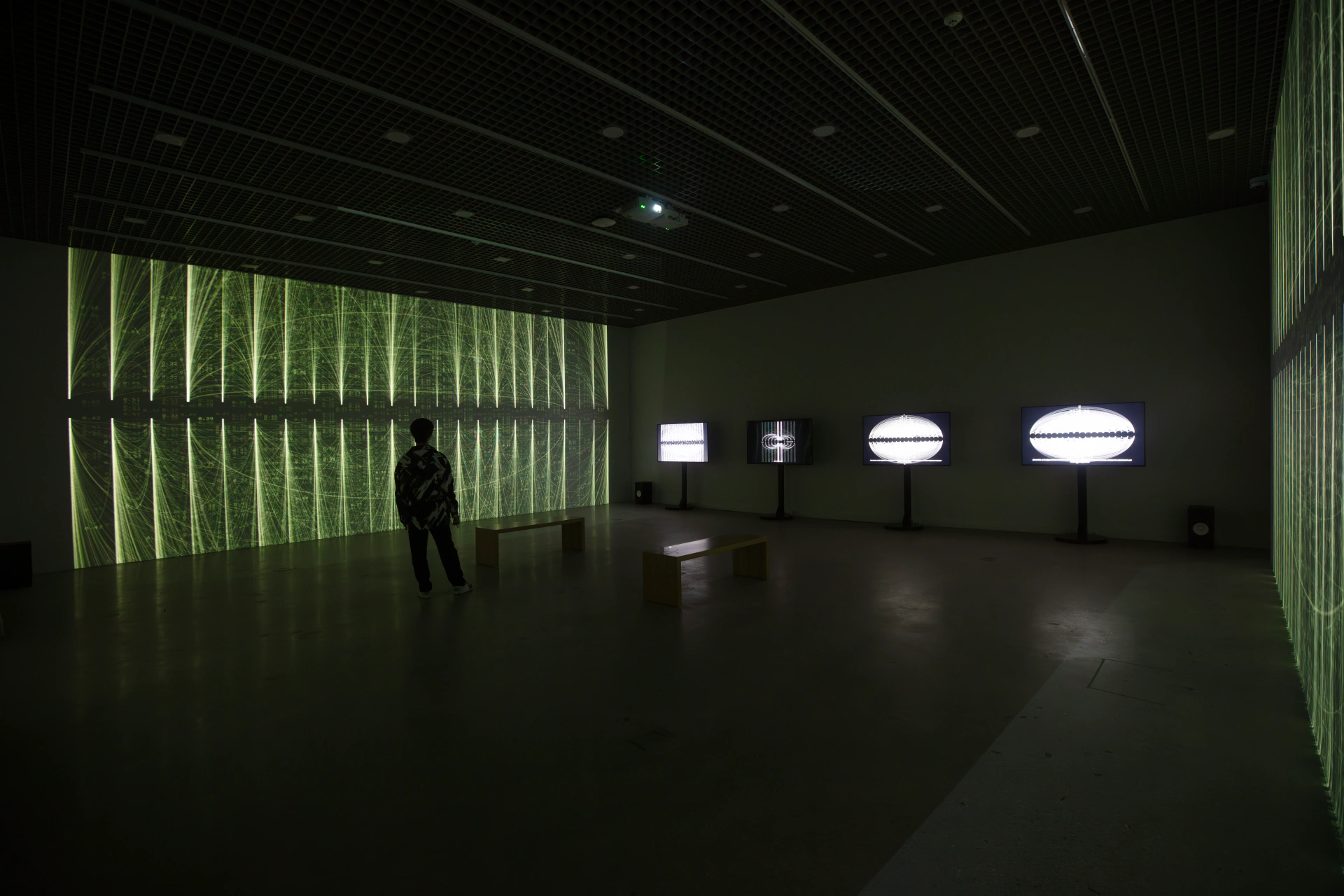 SoTA: visitor standing inside the gallery with green projection and four screens. Artwork by Jeanyoon Choi, Euan Kang, SeJoon Park, Minhyeok Seo, Prof. Yiyun Kang.